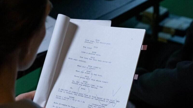 Person reading a script with notes in a dimly lit workspace, viewed over the shoulder.